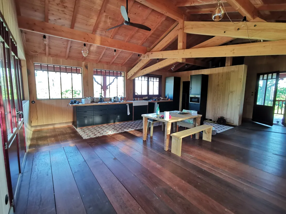Salle aménagée sous la charpente apparente, table en bois et cuisine en fond de pièce